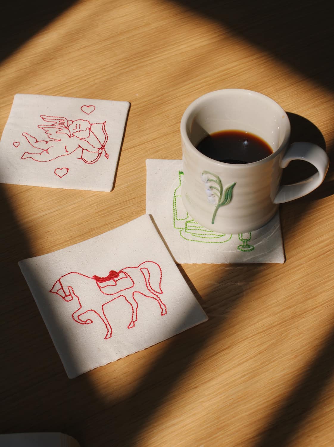 Handmade embroidered coaster set with horse and cupid motifs, artisan fabric coasters with ceramic mug on wooden table, aesthetic home styling for coffee and tea rituals
