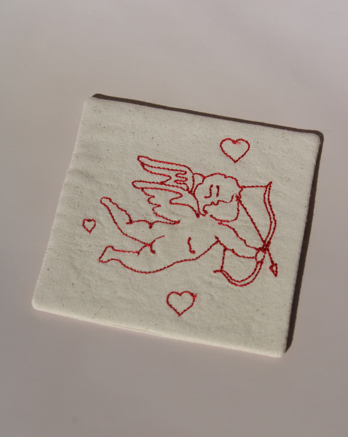 Amour Handsewn Embroidered Coaster with cupid and heart motif stitched in red on natural cotton handmade drink coaster.
