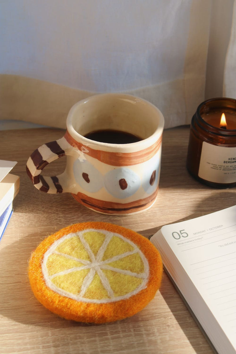 A felt orange slice coaster in the foreground on a wooden table, next to a ceramic mug filled with coffee, an open notebook, and a lit candle.