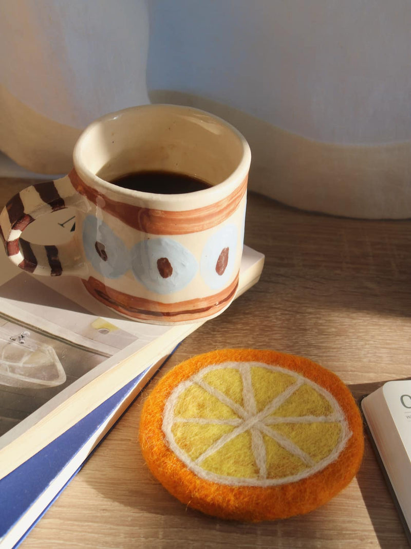 A felt orange slice coaster in the foreground on a wooden table, next to a ceramic mug filled with coffee, an open notebook, and a lit candle.