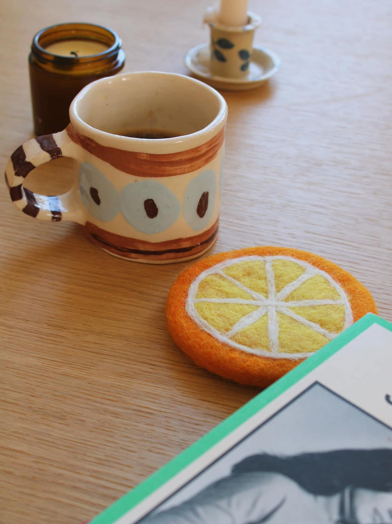 A felt orange slice coaster in the foreground on a wooden table, next to a ceramic mug filled with coffee, an open notebook, and a lit candle.