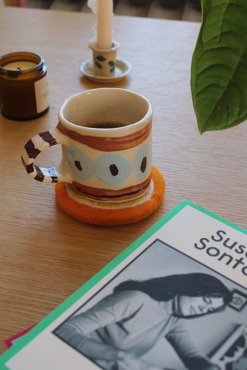A felt orange slice coaster in the foreground on a wooden table, next to a ceramic mug filled with coffee, an open notebook, and a lit candle.