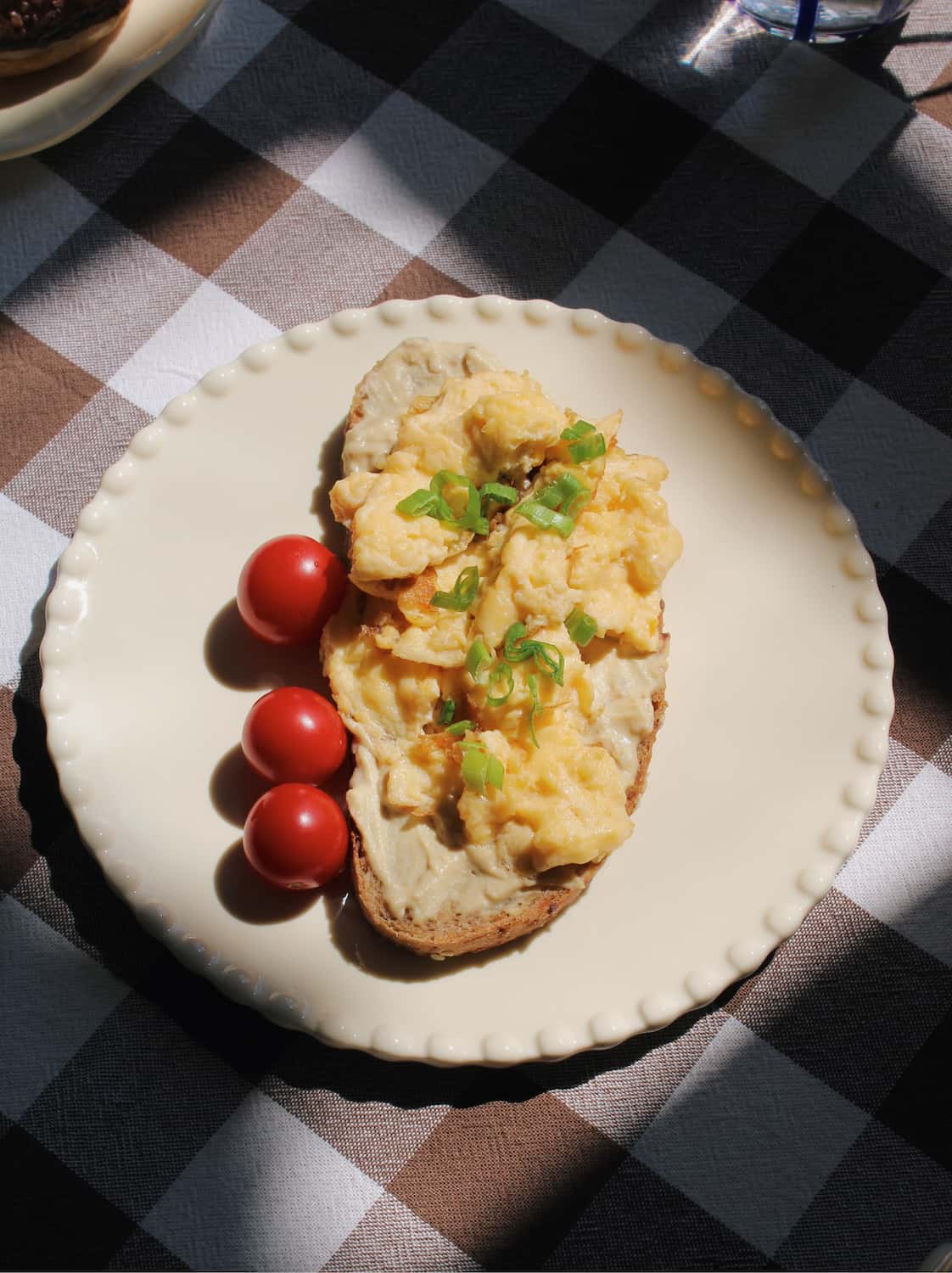 Scrambled eggs on toast with cherry tomatoes and green onions on a white plate, placed on a checkered tablecloth.