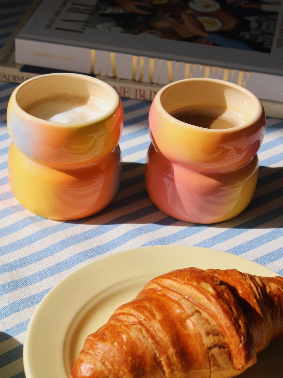 Two ceramic cups with a gradient design, one filled with coffee, on a striped tablecloth with a croissant.