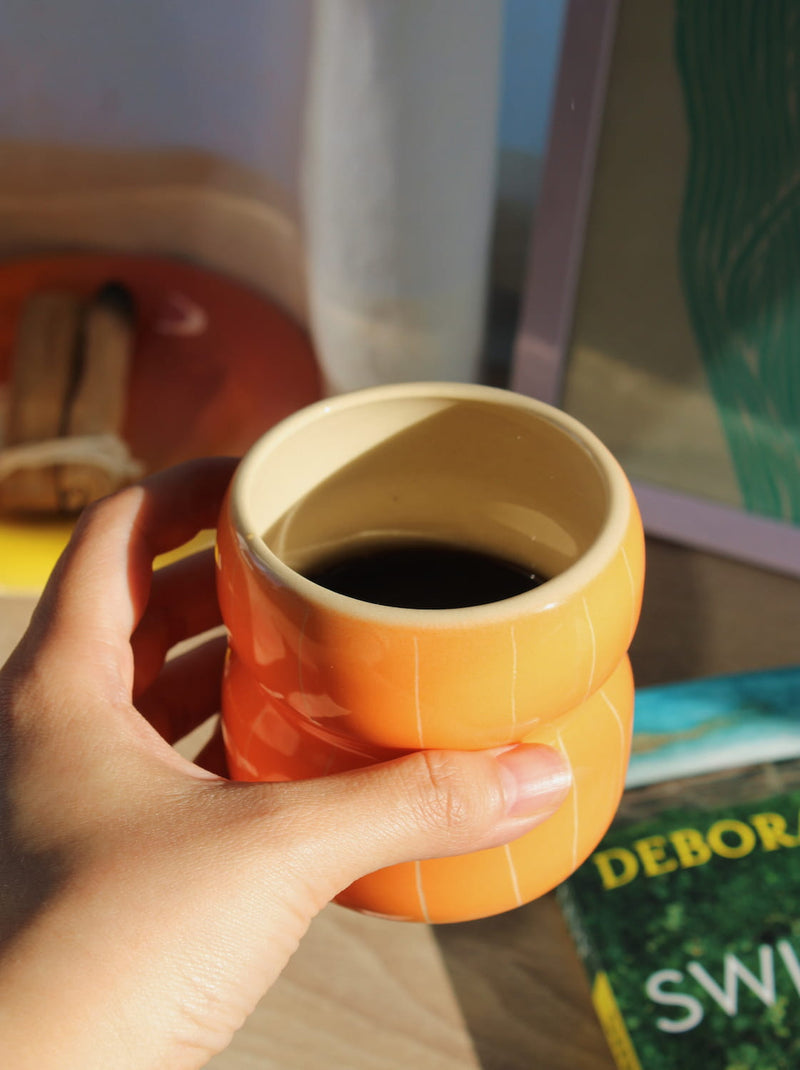 Belly Tangerine Ceramic Mug in glossy orange with ribbed belly shape, styled on a wooden surface with a white vase, book and the Belly Sunset Ceramic Plate with palo santo in warm natural light.