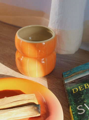 Belly Tangerine Ceramic Mug in glossy orange with ribbed belly shape, styled on a wooden surface with a white vase, book and the Belly Sunset Ceramic Plate with palo santo in warm natural light.
