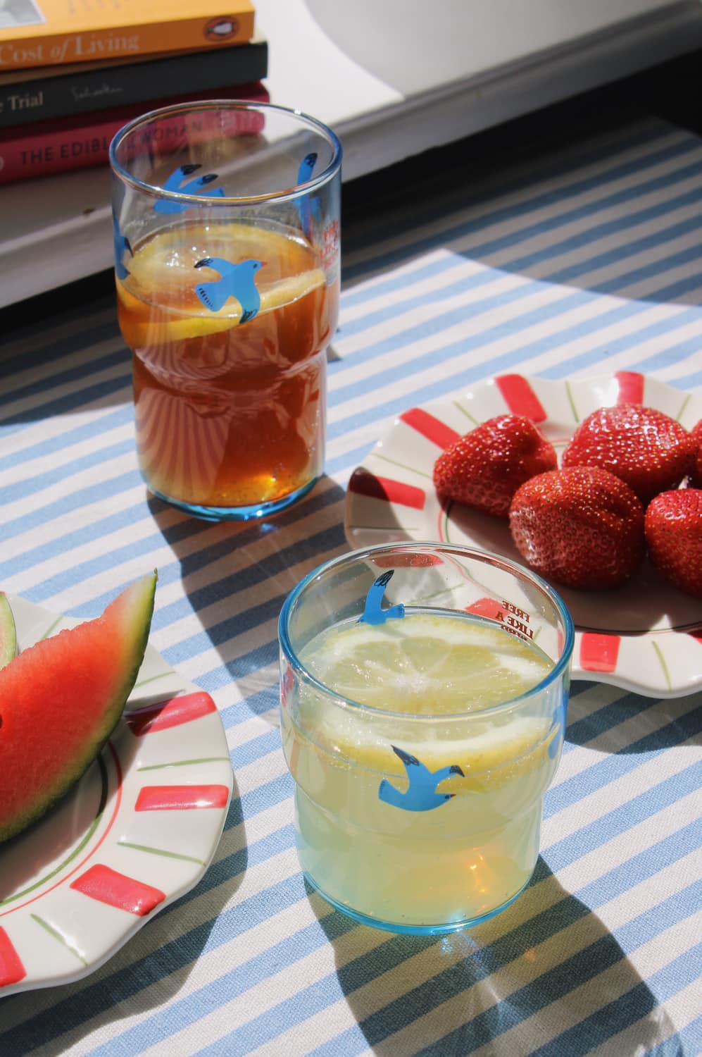 Two glasses of iced drinks with lemon slices on a striped tablecloth, accompanied by watermelon and strawberries.