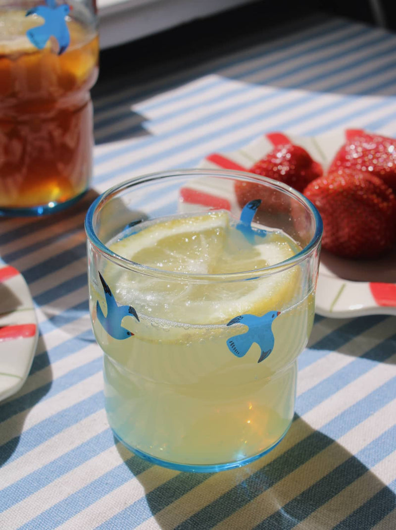 Glass of lemonade with a lemon slice and blue fish-shaped straw on a striped tablecloth.