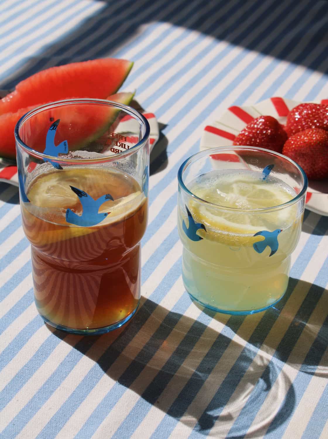 Two glasses of iced drinks on a striped tablecloth with watermelon and strawberries.