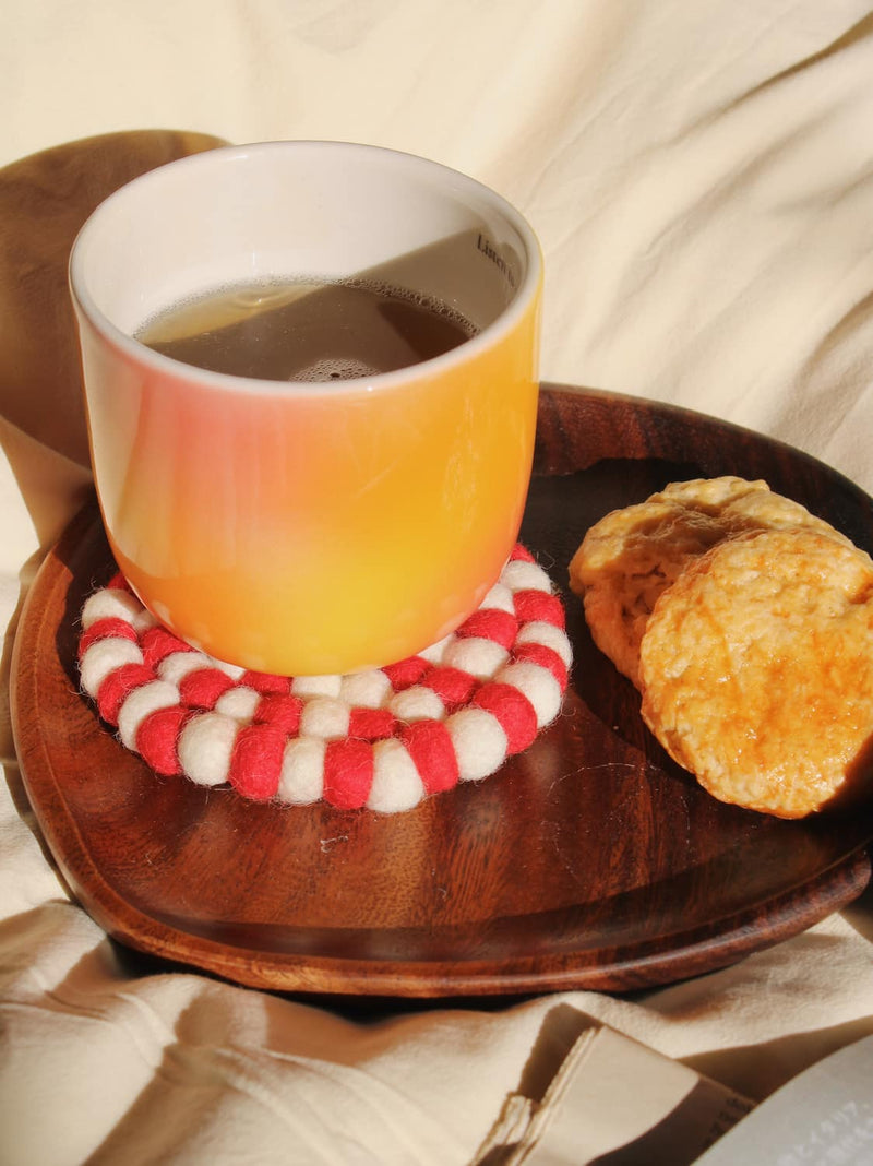 A ceramic mug with a warm orange-to-yellow gradient, filled with tea and resting on a red-and-white striped felt coaster, set on a dark wooden tray with two biscuits on a beige fabric background.