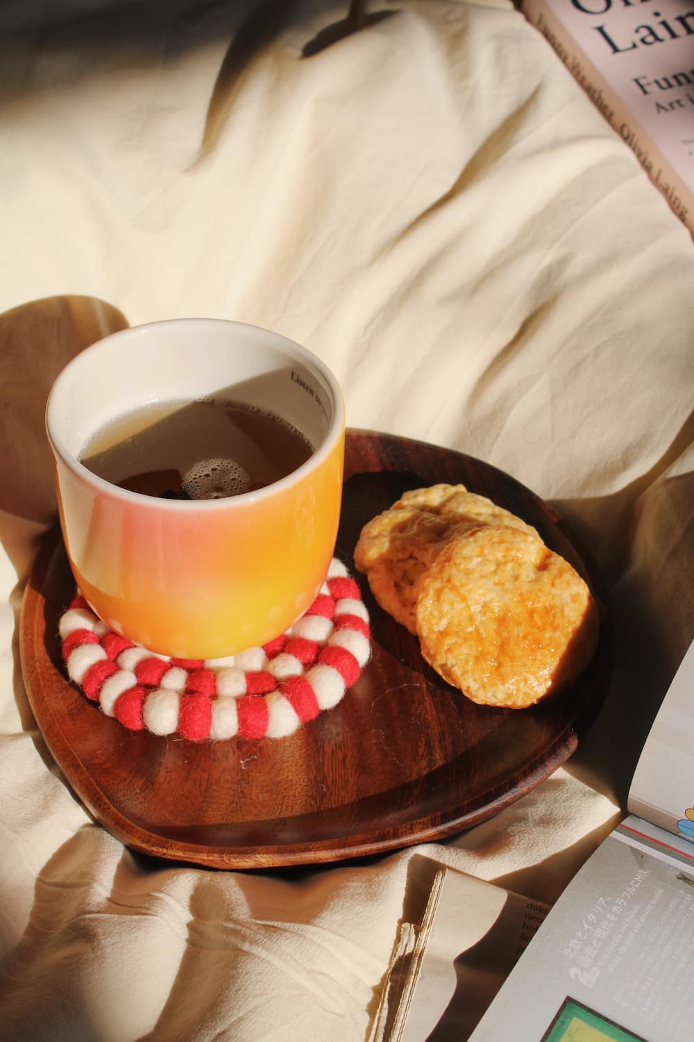 A ceramic mug with a warm orange-to-yellow gradient, filled with tea and resting on a red-and-white striped felt coaster, set on a dark wooden tray with two biscuits on a beige fabric background.