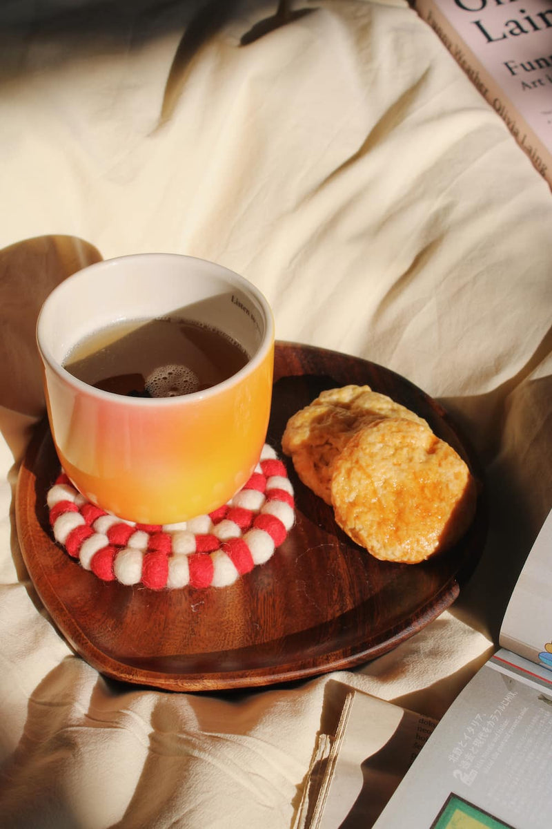 A ceramic mug with a warm orange-to-yellow gradient, filled with tea and resting on a red-and-white striped felt coaster, set on a dark wooden tray with two biscuits on a beige fabric background.