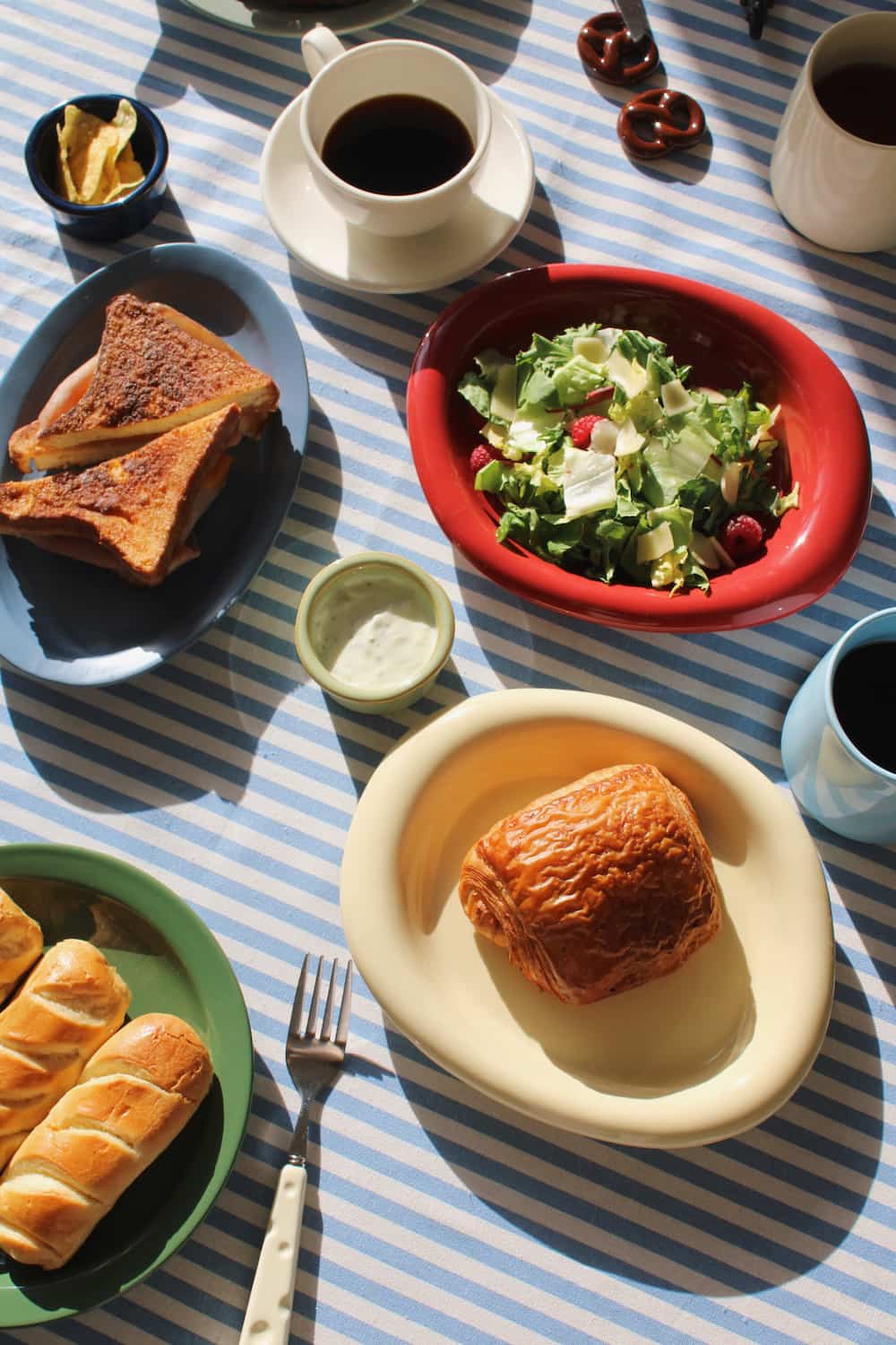 Colorful handmade ceramic tableware styled with breakfast food, coffee cups, plates, and bowls on a blue striped tablecloth