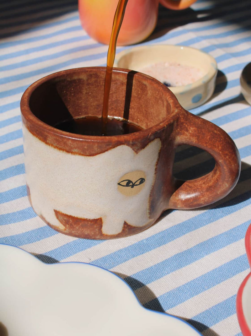 Ceramic mug with a cat design being filled with coffee on a striped tablecloth.