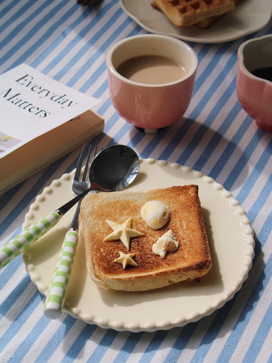 A styled breakfast scene on a blue and white striped tablecloth, featuring a plate of toasted bread topped with butter and shell decorations, a pink cup of coffee, and a book titled 'Everyday Matters'.