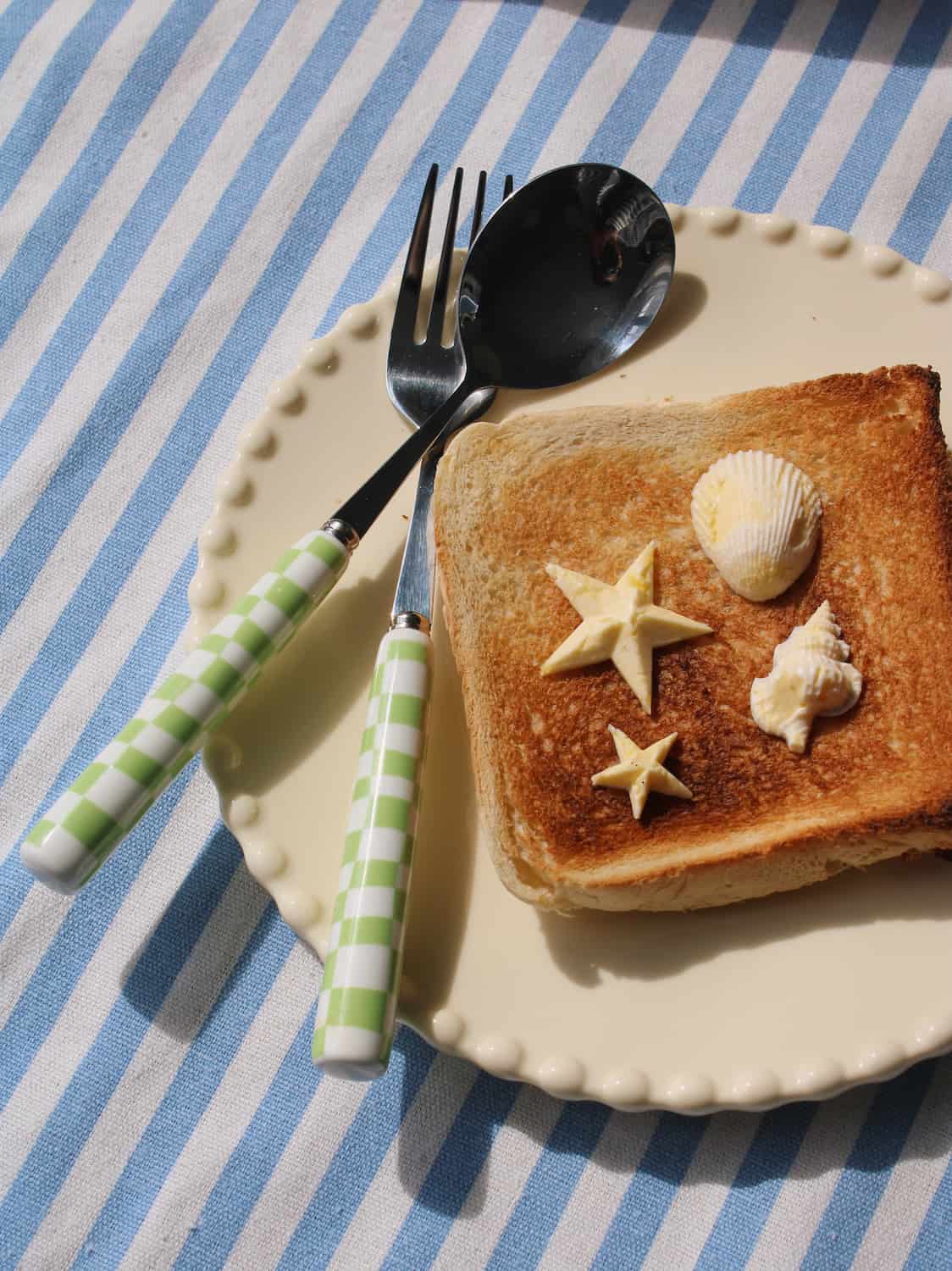 A styled tabletop scene with a beige scalloped plate holding a slice of toasted bread topped with butter shaped into stars and seashells, alongside a green-and-white checkered fork and spoon on a blue-and-white striped cloth.
