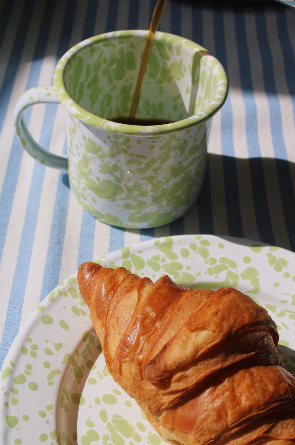 Green patterned clover enamel mug with coffee being poured next to a croissant on a striped tablecloth.