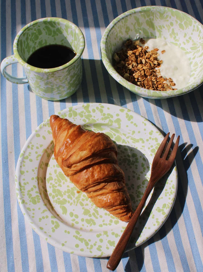 Breakfast setting with a croissant on a plate, coffee in a mug, and granola in a bowl on a striped tablecloth.