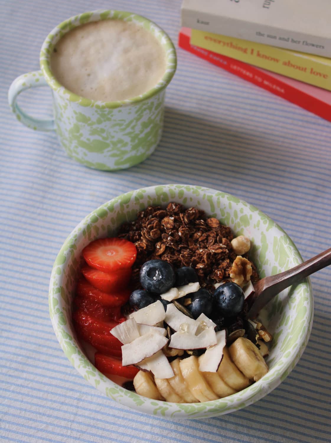 Enamel bowl of granola with fruits and an enamel mug of coffee on a striped surface
