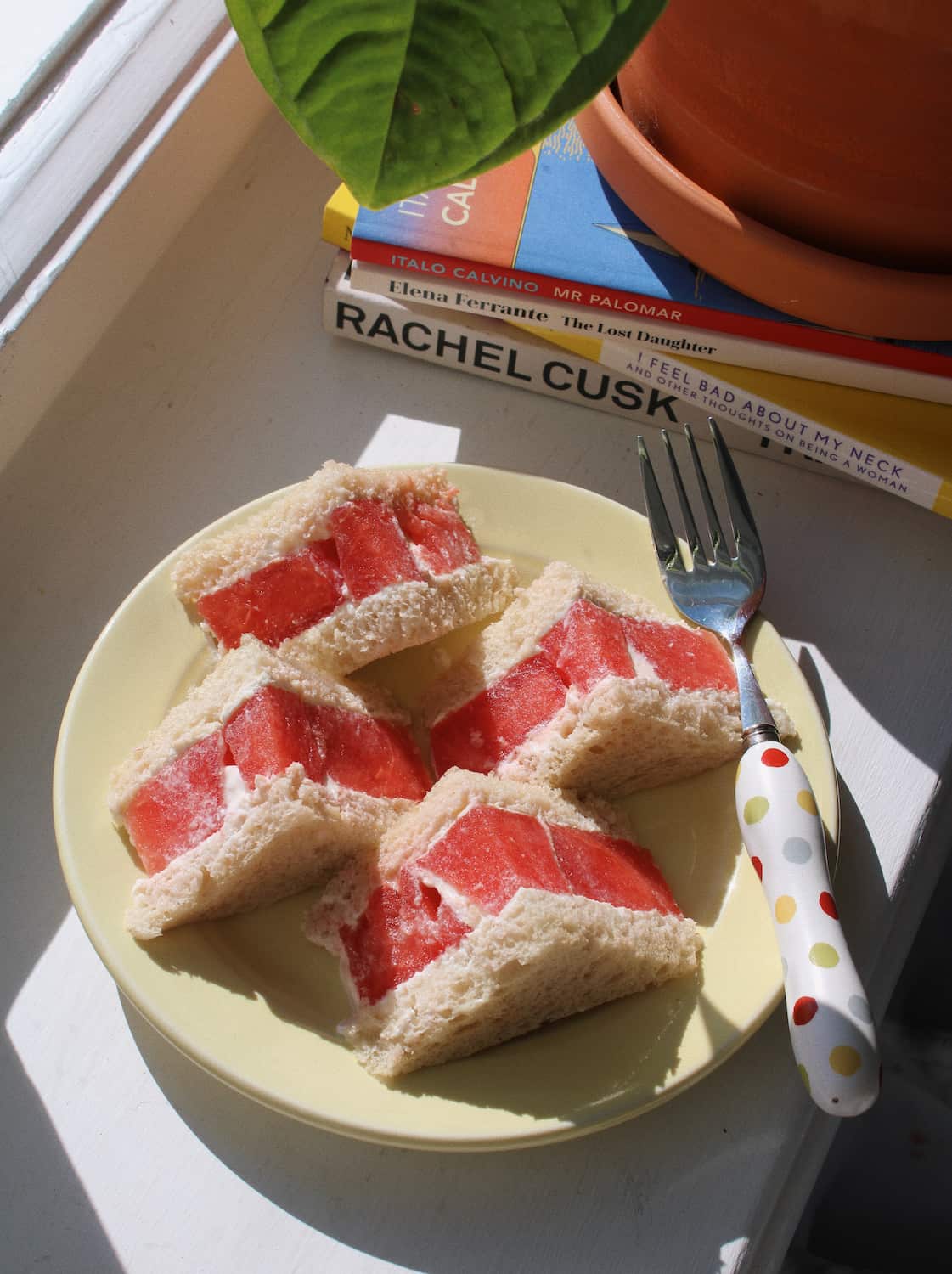 A plate of triangular sandwich slices with red filling, a fork with a polka-dot handle, and stacked books including one titled Rachel Cusk, placed on a windowsill with a plant leaf visible.