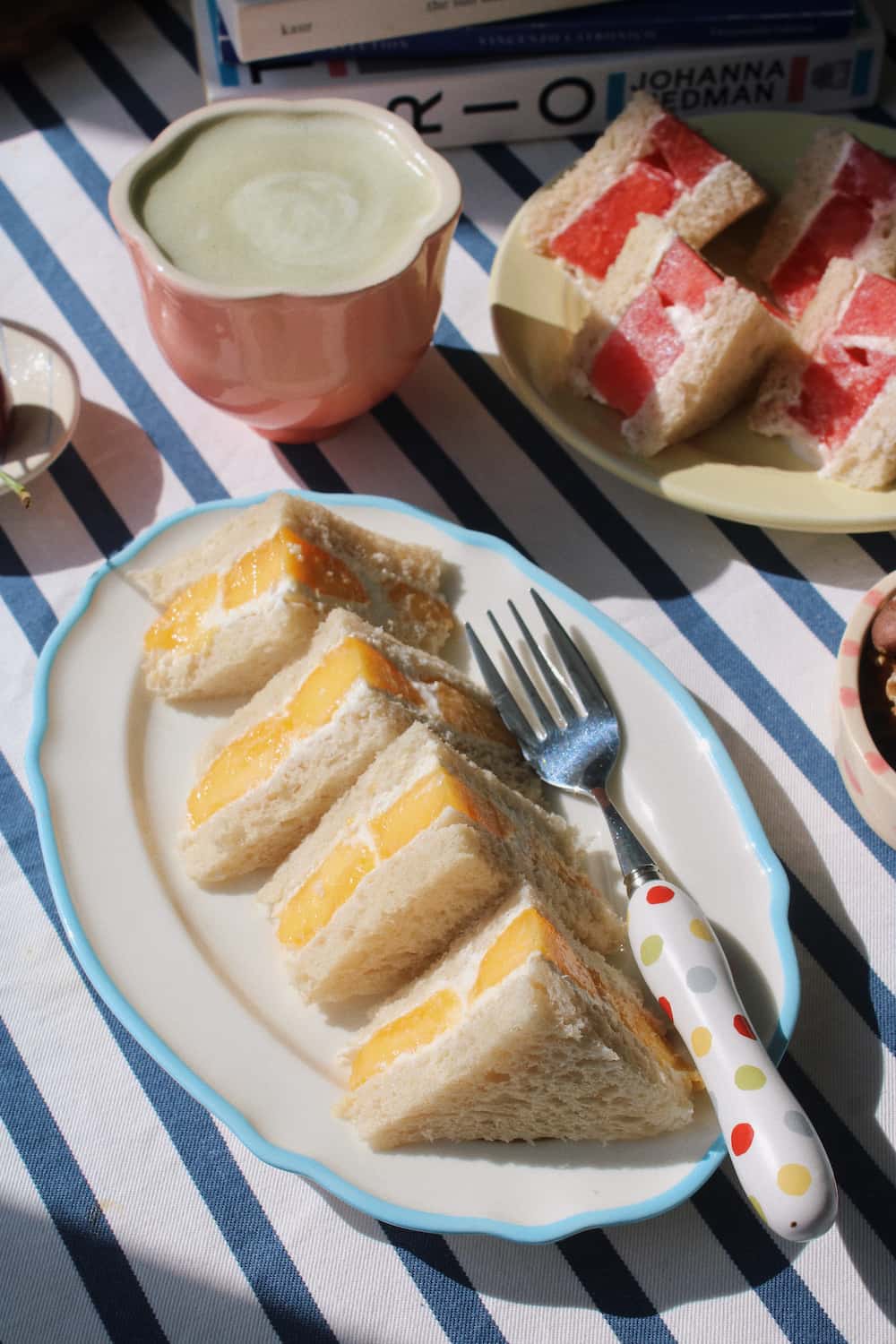 A close-up of a breakfast scene featuring sandwich slices with yellow and red fillings on a white plate with a blue rim, alongside a pink cup of iced coffee and another plate of sandwich slices on a blue-and-white striped tablecloth.