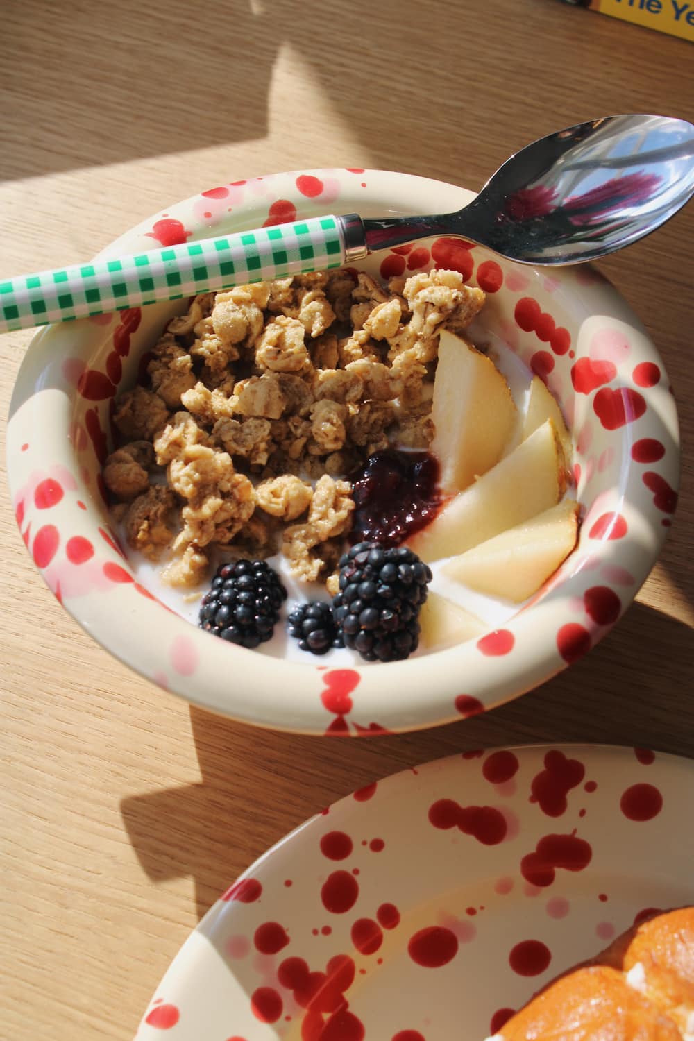 A close-up of a breakfast bowl on a wooden table, featuring granola, sliced apple, blackberries, and a dark berry compote in a white bowl with red speckles. A green-and-white checkered spoon handle rests across the bowl.