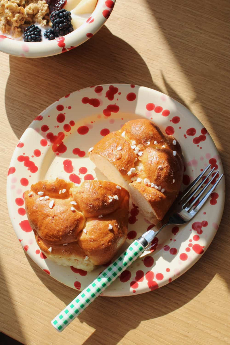 A close-up overhead shot of a white ceramic plate with red speckles, holding two braided bread rolls topped with coarse salt, on a wooden table. A green-and-white checkered fork rests on the plate, and part of a matching bowl is visible at the top edge.