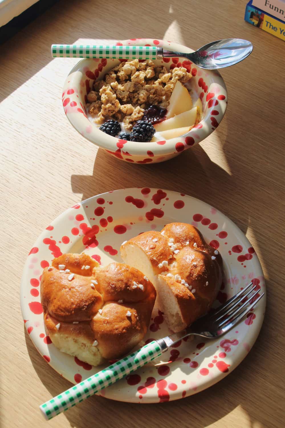 A breakfast scene on a wooden table with a bowl of granola and fruit in the background and a plate with bread rolls in the foreground. The plate and spoon have a white base with red polka dots, and the spoon handle is green and white checkered.