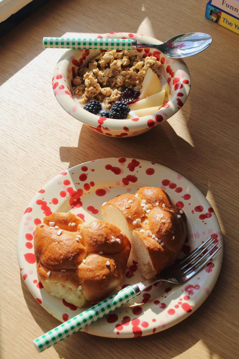 A breakfast scene on a wooden table with a bowl of granola and fruit in the background and a plate with bread rolls in the foreground. The plate and spoon have a white base with red polka dots, and the spoon handle is green and white checkered.