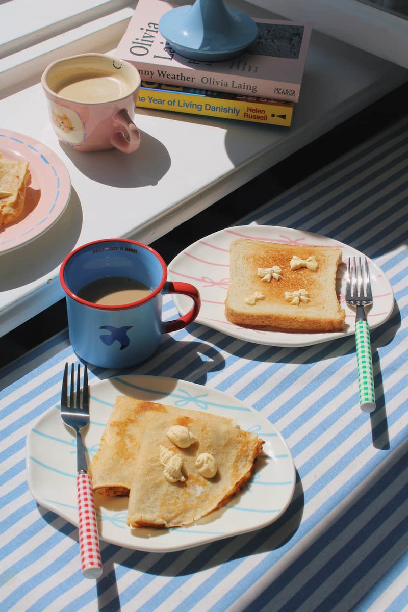 A styled breakfast scene with two white plates holding folded crepes topped with butter, a blue mug of coffee with a red rim, and a plate of toast with butter on a striped blue-and-white tablecloth in bright sunlight.