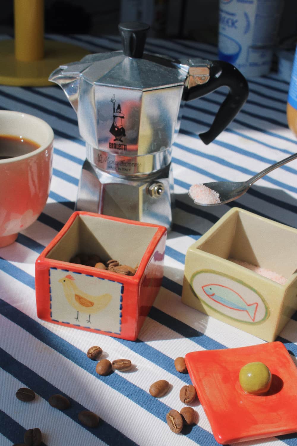 A styled coffee scene on a blue-and-white striped tablecloth with a silver moka pot, a cup of coffee, coffee beans, and two decorative ceramic containers holding pink sugar and a green olive.