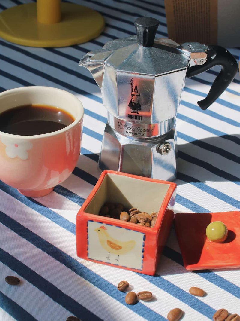 A silver aluminum moka pot sits on a blue-and-white striped tablecloth beside a pink cup of coffee, a small square orange container filled with coffee beans, and its lid with a green grape.