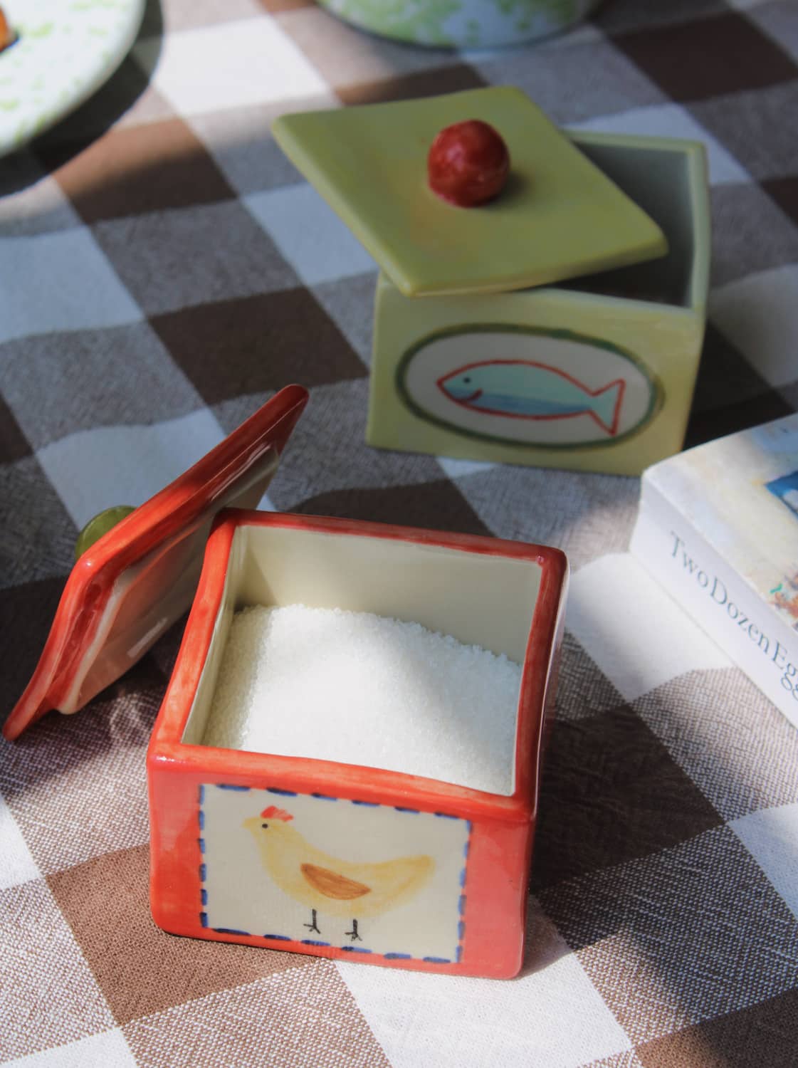 Sardina Ceramic Box in pale yellow and Henley Trinket Box in red with hand-painted chicken illustration, styled open together on a brown gingham surface with a Bialetti moka pot and stacked books in the background.
