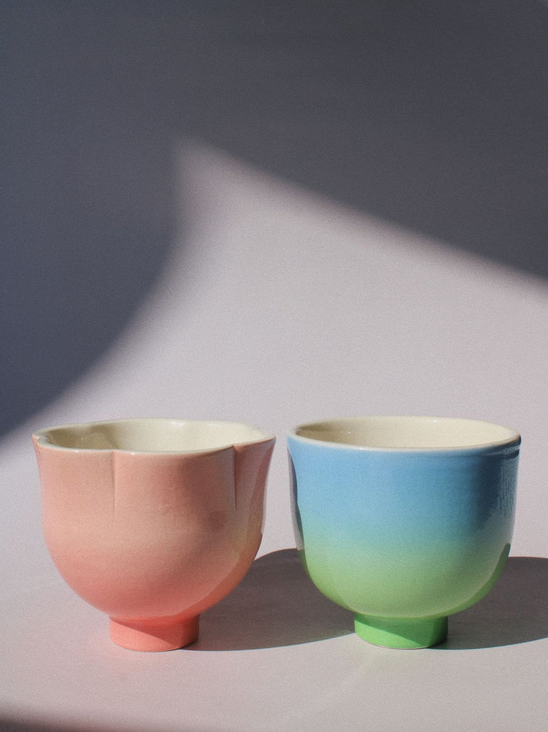 Two handmade ceramic cups from the Yunomi collection side by side, on a soft pink surface.