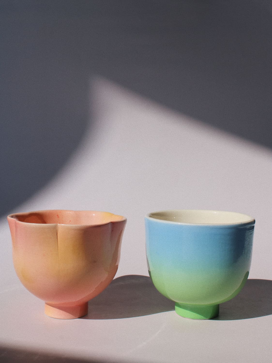 Two handmade ceramic cups with a pastel ombre finish, one in peach and yellow tones and the other in blue, green, and yellow tones, set against a gray background with dramatic shadows.