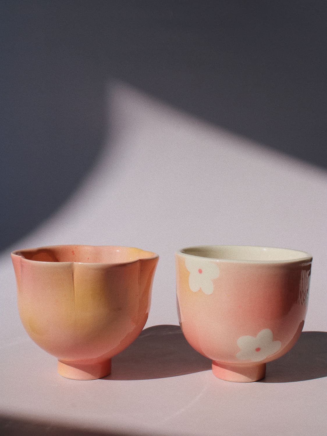 Two small handmade ceramic cups in a soft peach-pink glaze, one with a scalloped rim and the other decorated with white cherry blossom motifs, set against a dark gray background with dramatic shadow play.