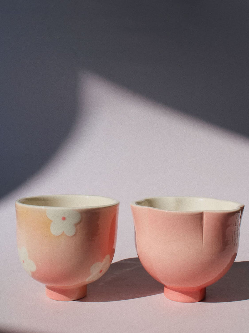 Two small pink ceramic cups with white floral patterns, set against a dark gray background with dramatic diagonal shadows.