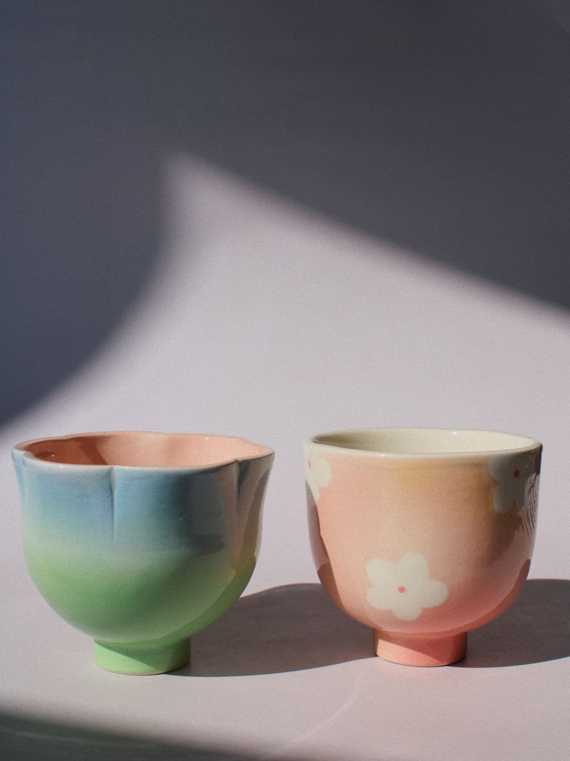 Two pastel ceramic cups with a soft gradient finish, one transitioning from blue to green, and the other from peach to pink with a white flower motif, set against a gray background with dramatic shadows.