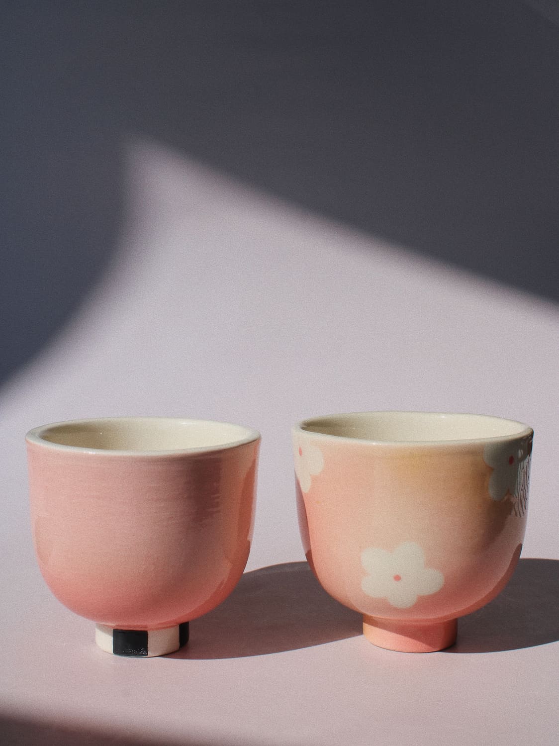 Two small pink ceramic cups with a glossy finish, one solid pink and the other with a pink-to-cream gradient and a white cherry blossom motif, set against a soft pink background with dramatic shadows.
