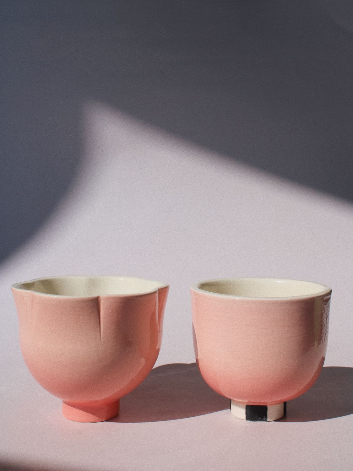 Two small pink ceramic cups with a matte finish and cream-colored interiors, set against a soft pink background with dramatic diagonal shadows.
