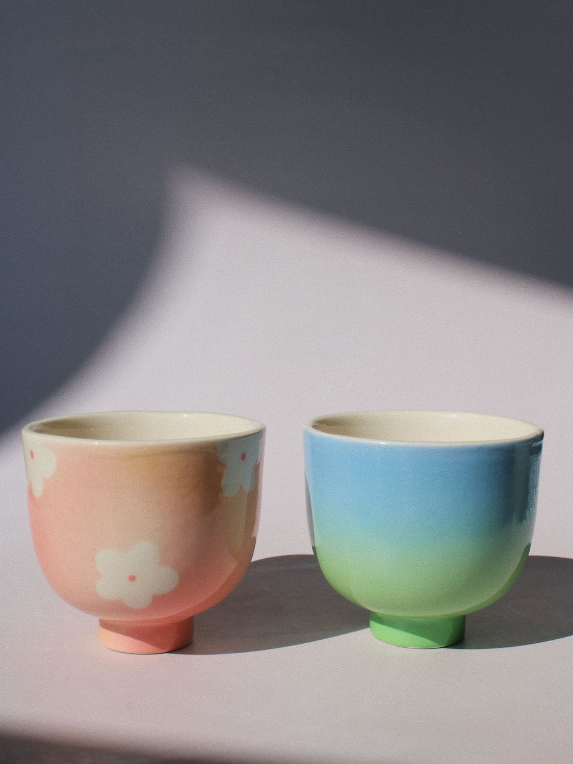 Two handmade ceramic cups from the Yunomi collection side by side, on a soft pink surface.