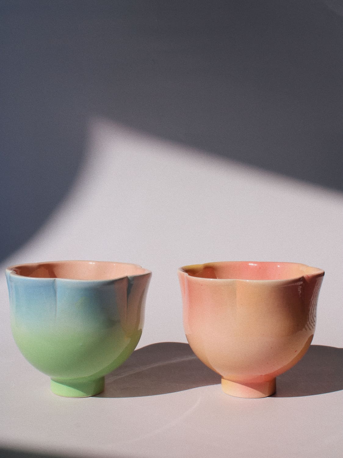 Two pastel ceramic cups with a soft gradient finish, one transitioning from blue to green, and the other from pink to peach, set on a white surface with a gray background and a soft shadow.