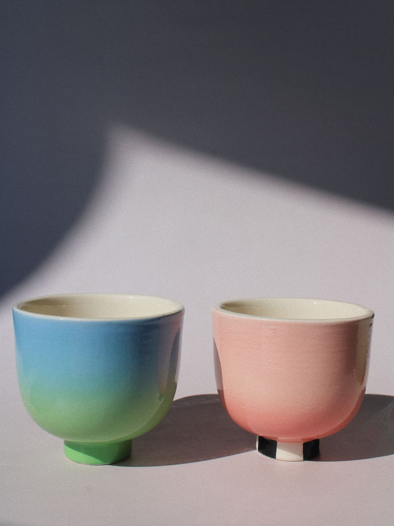 Two handmade ceramic cups from the Yunomi collection side by side against a pink background