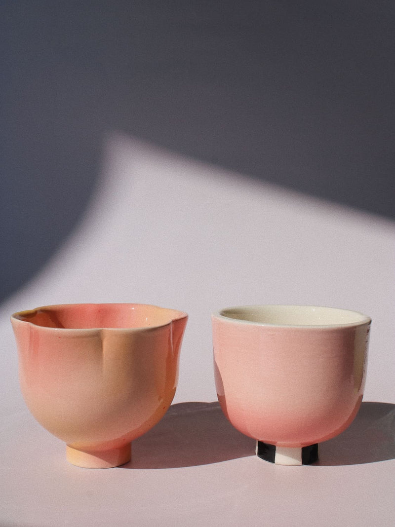 Two small ceramic cups with a soft pink and peach gradient glaze, one with a flared rim and the other with a simple rounded shape, set against a dark gray background with a light pink surface and dramatic shadows.