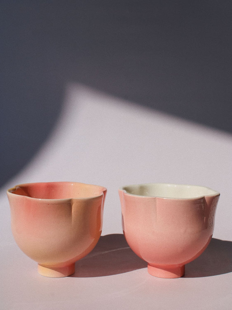 Two small ceramic cups with a soft pink and peach gradient glaze, one with a pale peach exterior and one with a pale cream interior, set against a dark gray background with a light-colored surface and dramatic shadows.