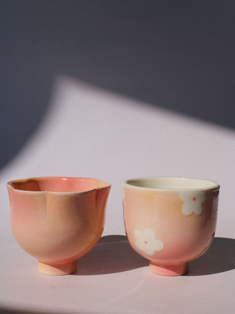 Two small ceramic cups with a soft pink and peach gradient glaze, one plain and one decorated with white cherry blossom motifs, set against a dark gray background with dramatic diagonal shadows.