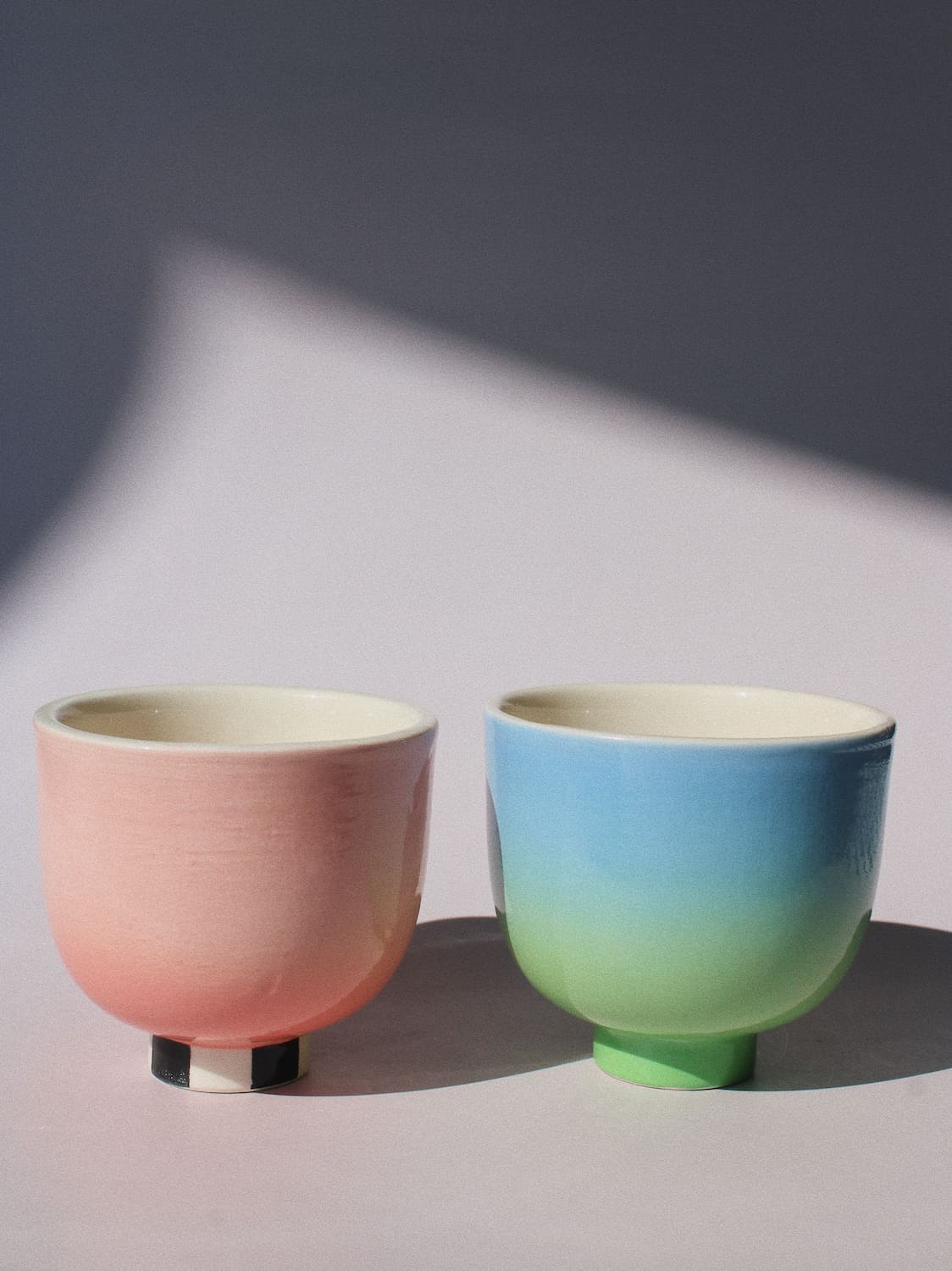 Two handmade ceramic cups from the Yunomi collection side by side, on a soft pink surface.