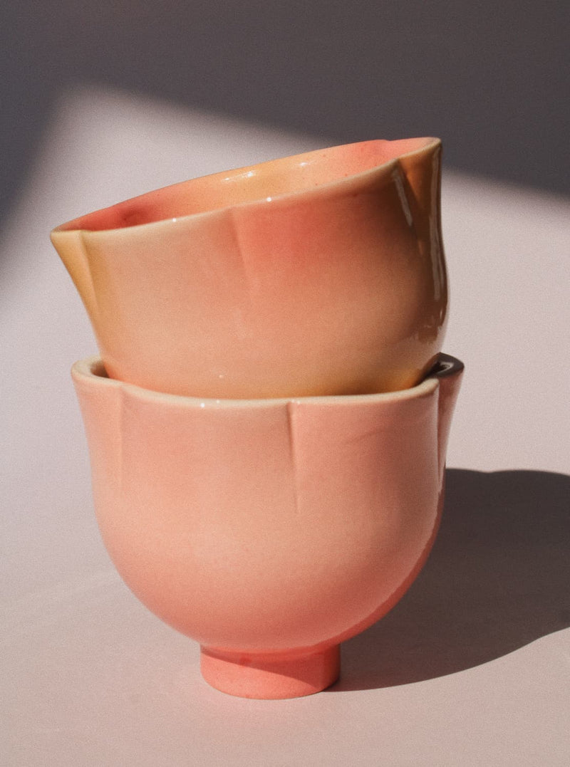 Two ceramic cups stacked together, featuring a glossy gradient glaze that transitions from a warm peach-pink at the bottom to a soft cream-beige at the rim, set against a neutral background with dramatic diagonal shadows.