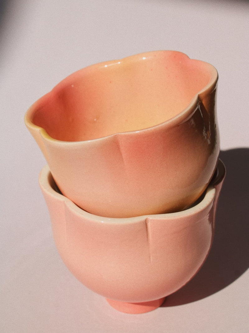Two stacked ceramic cups with a scalloped rim, featuring a soft peach to pale pink gradient glaze, set against a plain light background.