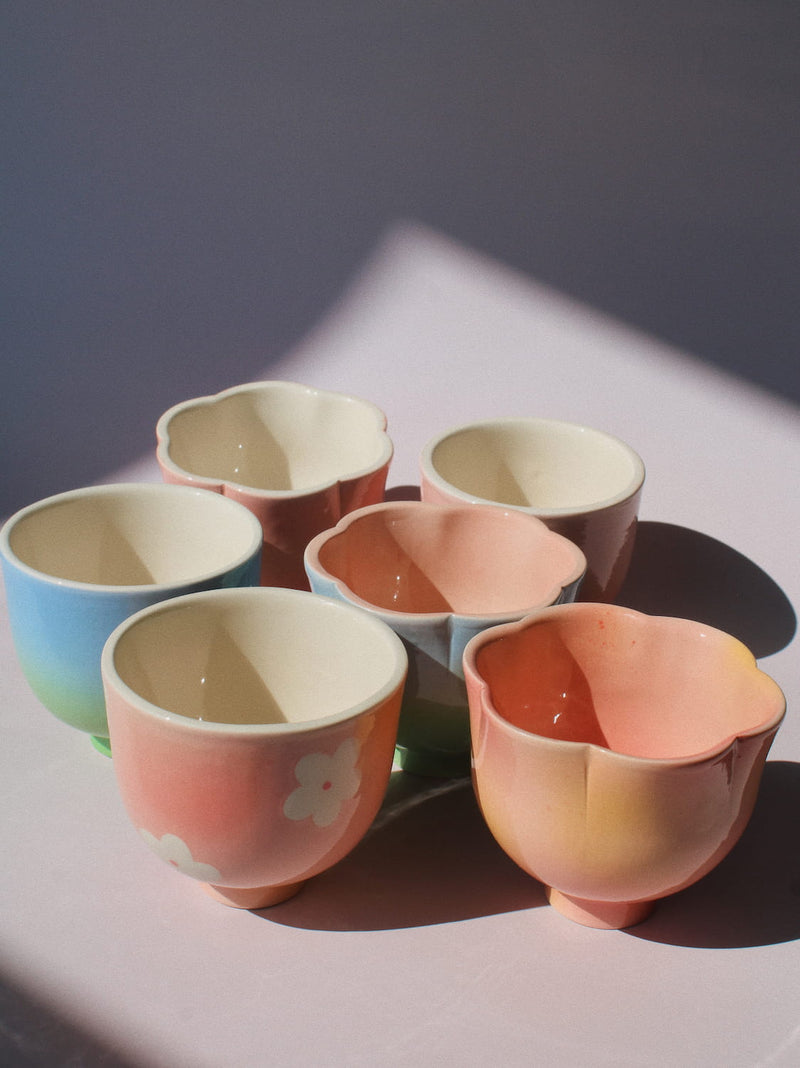 Six pastel ceramic teacups with scalloped flower-like rims, arranged in a cluster on a pink surface under direct sunlight. The cups feature soft gradients of pink, peach, blue, and yellow, with one cup decorated with a white cloud motif.